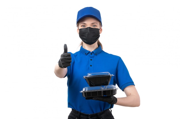 A front view young female courier in blue uniform black gloves and black mask holding food delivery bowls