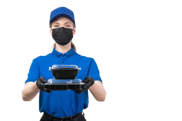 A front view young female courier in blue uniform black gloves and black mask holding food delivery bowls