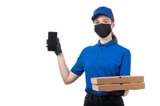 A front view young female courier in blue uniform black gloves and black mask holding food delivery packages and smartphone