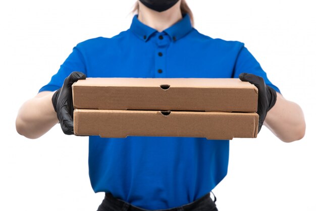 A front view young female courier in blue uniform black gloves and black mask holding food delivery packages