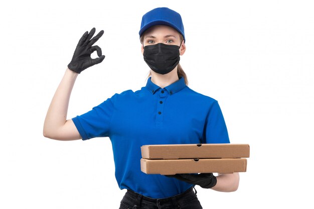 A front view young female courier in blue uniform black gloves and black mask holding food delivery packages