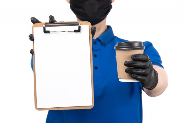 A front view young female courier in blue uniform black mask and gloves holding coffee cup and notepad