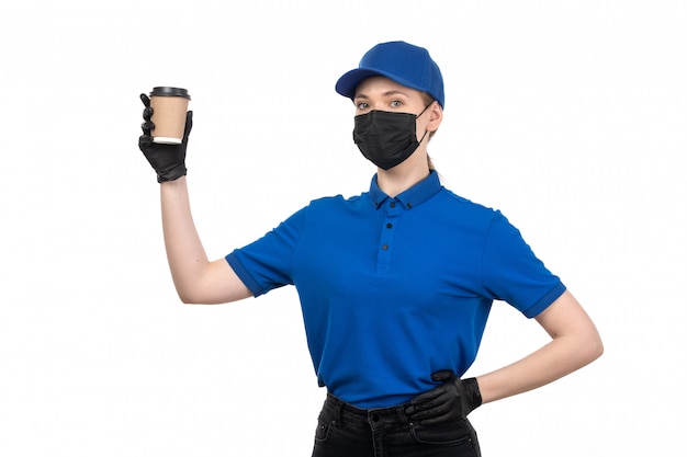 A front view young female courier in blue uniform black mask and gloves holding coffee cup