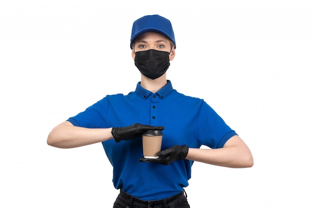Молодая женщина-курьер в синей униформе, черной маске и перчатках, держащая чашку кофе, вид спереди