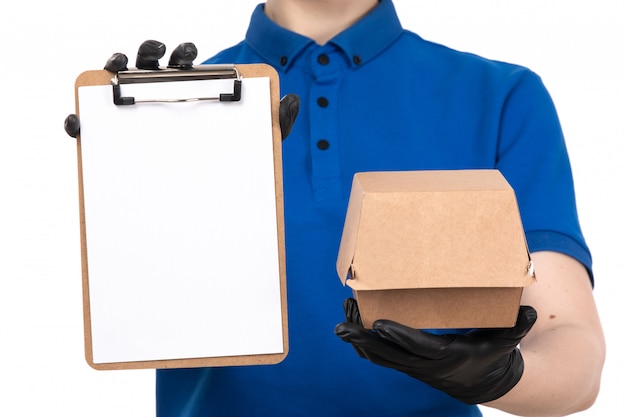 A front view young female courier in blue uniform black mask and gloves holding food delivery package and notepad