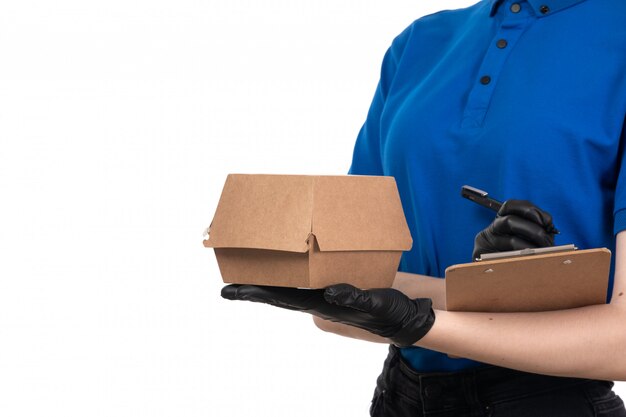 A front view young female courier in blue uniform black mask and gloves holding food delivery package and notepad