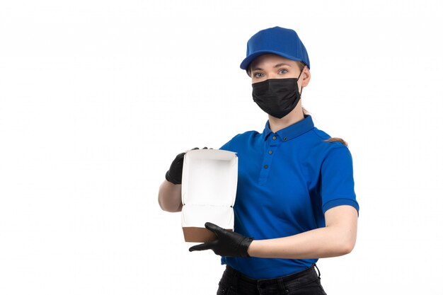 A front view young female courier in blue uniform black mask and gloves holding food delivery package