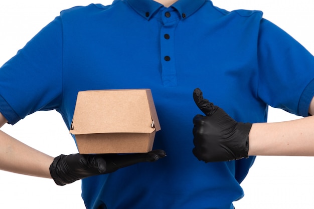 A front view young female courier in blue uniform black mask and gloves holding food delivery package