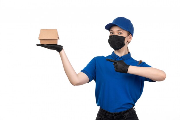 A front view young female courier in blue uniform black mask and gloves holding food delivery package