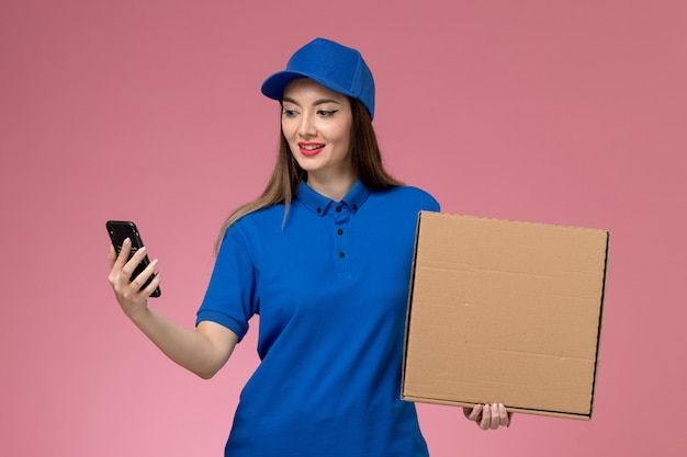 Молодая женщина-курьер в синей форме и плаще, держащая пустой ящик для еды и телефон на розовой стене, вид спереди