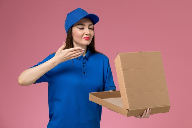 Front view young female courier in blue uniform and cape holding empty food box smelling it on pink wall 