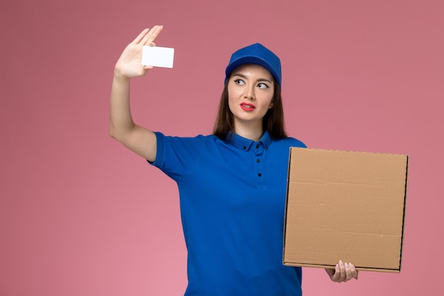 Молодая женщина-курьер в синей форме и плаще, держащая коробку с едой и карточку на розовой стене, вид спереди