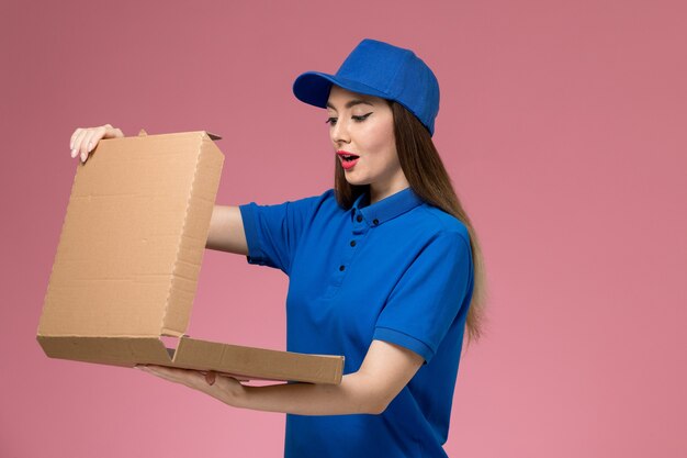 Front view young female courier in blue uniform and cape holding food box opening it on pink desk 