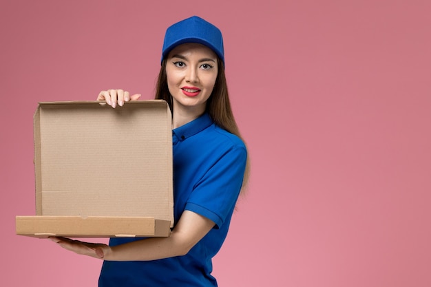 Front view young female courier in blue uniform and cape holding food box and opening it on pink wall 