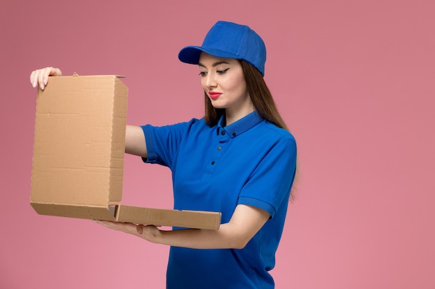 Front view young female courier in blue uniform and cape holding food box opening it on pink wall 