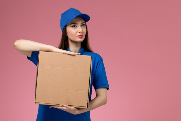 青い制服と淡いピンクの壁の配達制服の仕事で食品配達ボックスを保持している岬の正面図若い女性の宅配便