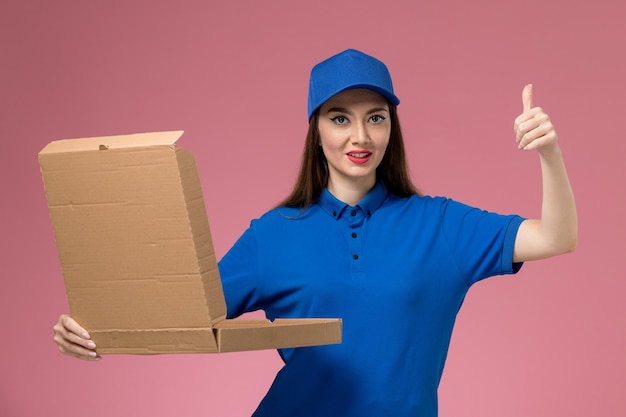 Front view young female courier in blue uniform and cape holding food delivery box opening it on light pink wall 
