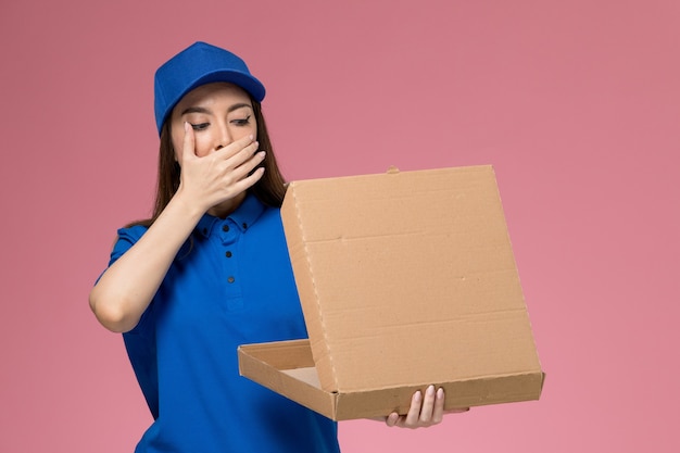 Front view young female courier in blue uniform and cape holding food delivery box and opening it on pink desk 