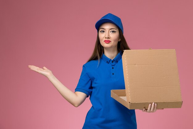 Вид спереди молодая женщина-курьер в синей форме и плаще держит коробку для доставки еды, открывающую ее на розовой стене