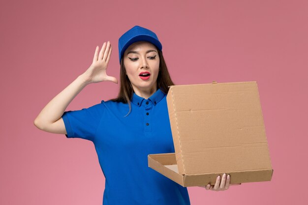 Front view young female courier in blue uniform and cape holding food delivery box opening it on the pink wall 