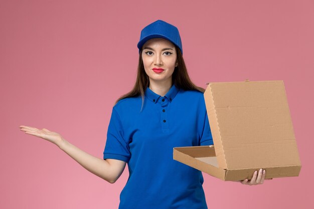 Front view young female courier in blue uniform and cape holding food delivery box opening it smiling on the pink wall 