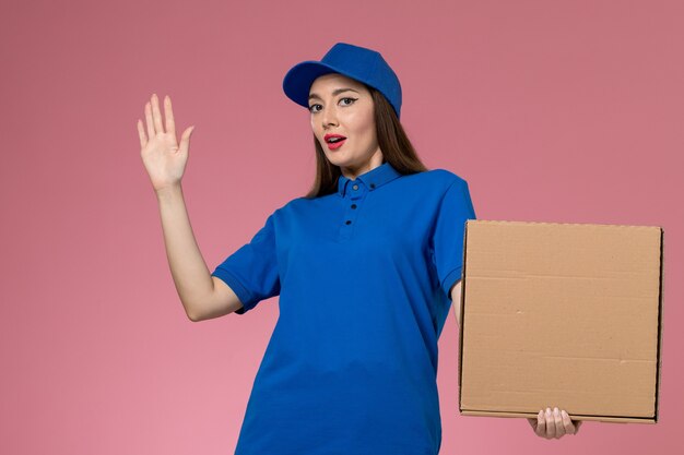 Front view young female courier in blue uniform and cape holding food delivery box on the pink wall