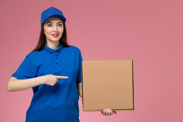 Молодая женщина-курьер в синей форме и плаще, держащая коробку для доставки еды на розовой стене, вид спереди