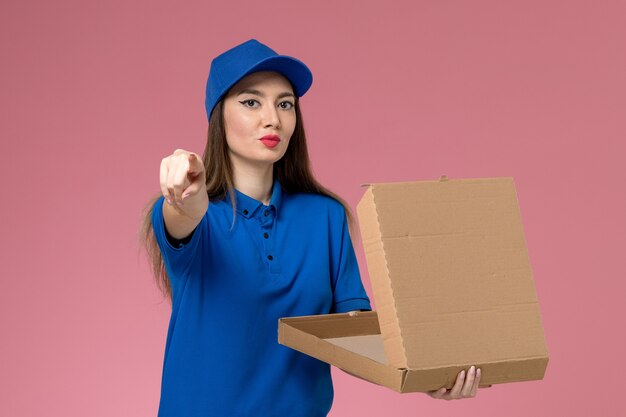 Front view young female courier in blue uniform and cape holding food delivery box posing on the pink wall 