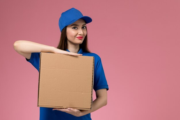 Вид спереди молодая женщина-курьер в синей форме и плаще, держащая коробку для доставки еды, улыбаясь на светло-розовой стене