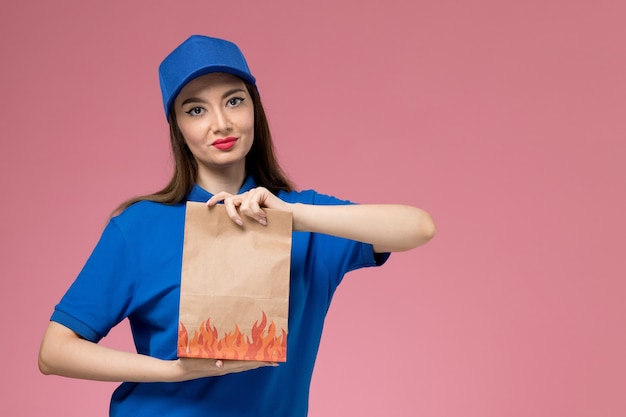淡いピンクの壁の労働者に紙の食品パッケージを保持している青い制服と岬の正面図若い女性の宅配便