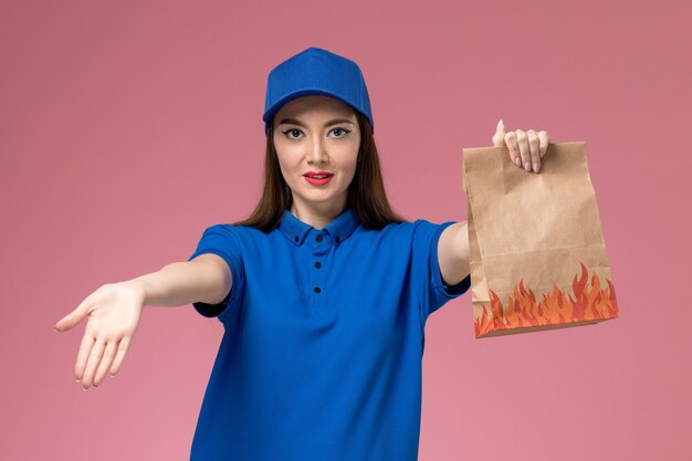 Front view young female courier in blue uniform and cape holding paper food package on light-pink wall 