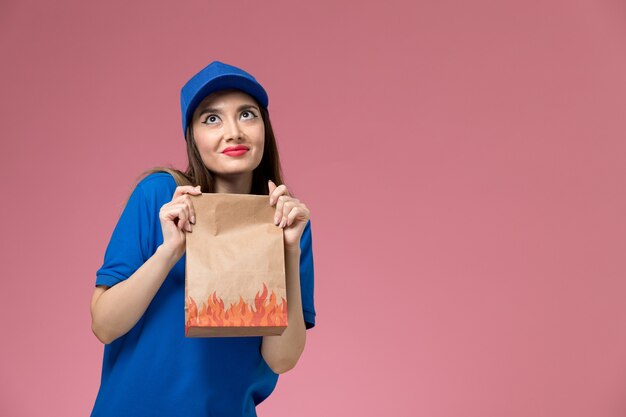 Front view young female courier in blue uniform and cape holding paper food package on the light pink wall 