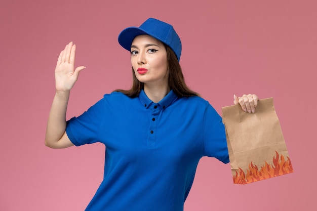 Front view young female courier in blue uniform and cape holding paper food package on the pink wall model