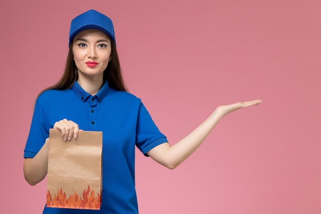 Front view young female courier in blue uniform and cape holding paper food package on pink wall