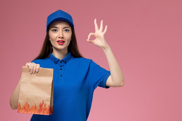 Front view young female courier in blue uniform and cape holding paper food package on the pink wall 