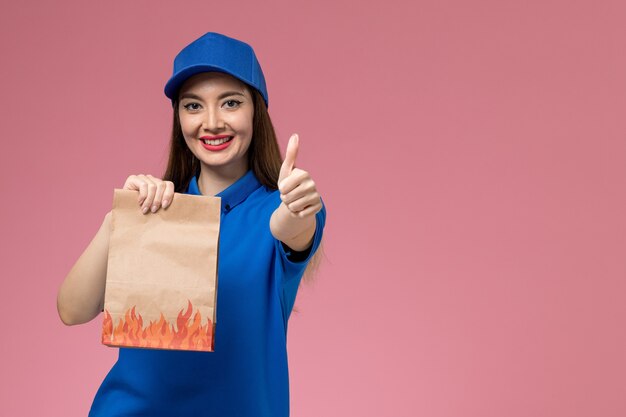 Front view young female courier in blue uniform and cape holding paper food package with smile on pink wall 