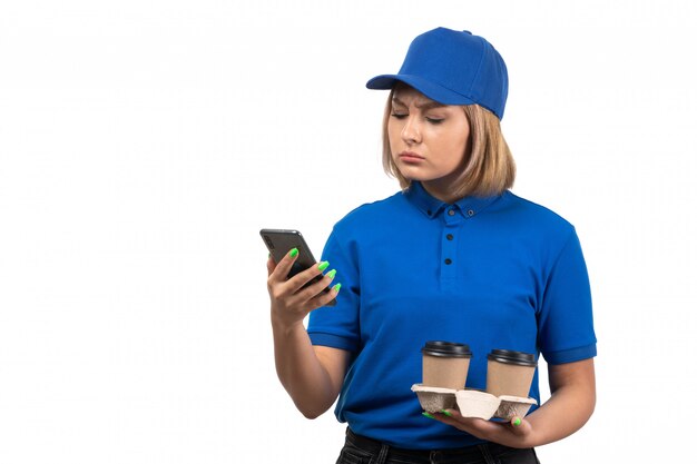 A front view young female courier in blue uniform holding phone and coffee cups