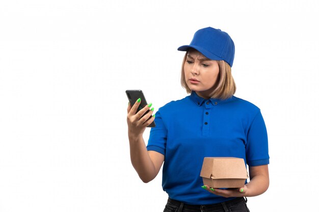 A front view young female courier in blue uniform holding phone and food delivery package