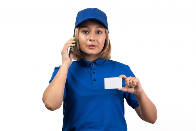 A front view young female courier in blue uniform holding phone and white card talking