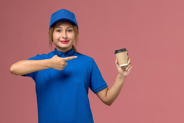 ピンクの壁に茶色のコーヒーの配達カップを保持している青い制服ポーズの正面図若い女性の宅配便、サービス制服配達女性の仕事