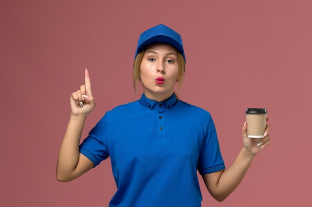 Giovane corriere femminile di vista frontale in uniforme blu che posa che tiene la tazza di caffè marrone di consegna, foto dell'operaio di lavoro della donna di consegna dell'uniforme di servizio