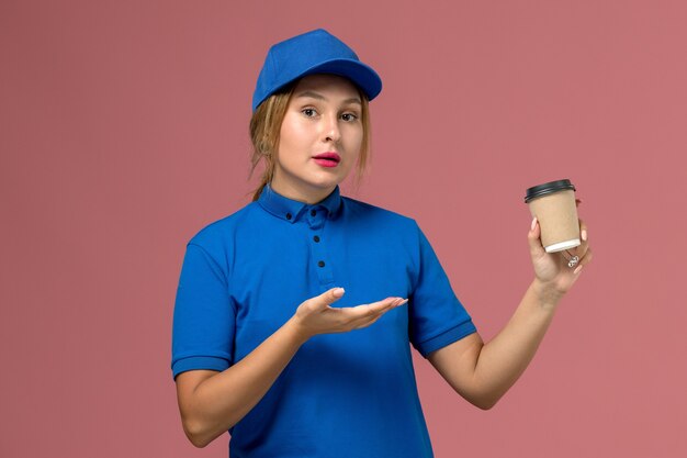 Front view young female courier in blue uniform posing holding brown delivery cup of coffee, service uniform delivery woman job
