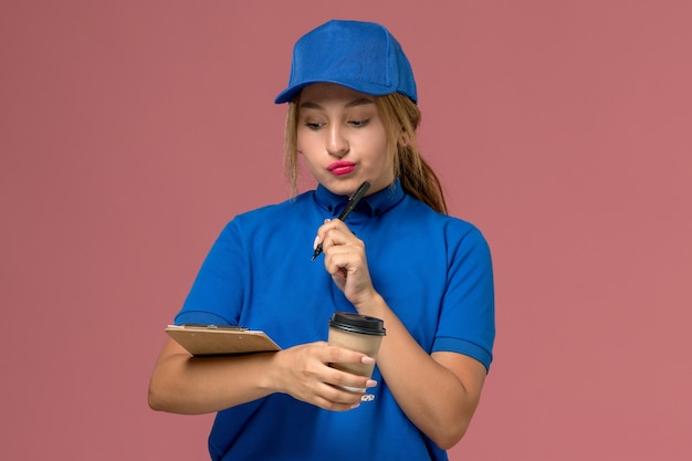 Front view young female courier in blue uniform posing holding cup of coffee and notepad thinking, service uniform delivery woman job