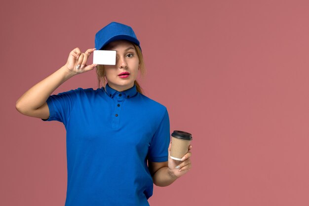 Front view young female courier in blue uniform posing holding cup of coffee and white card, service uniform delivery woman job worker