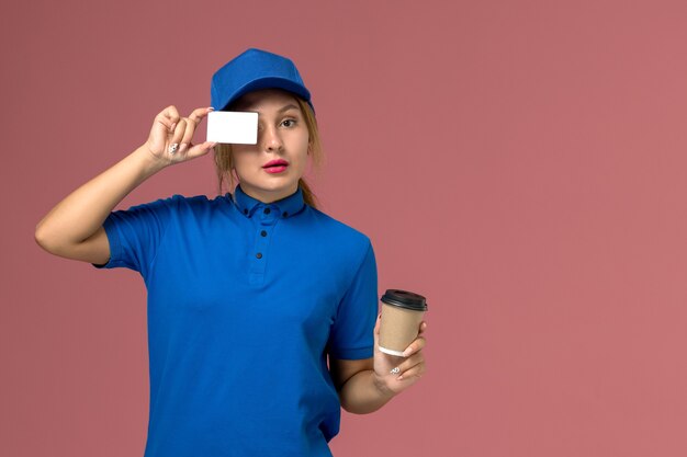 Вид спереди молодая женщина-курьер в синей форме позирует, держа чашку кофе и белую карточку, работница службы доставки униформы