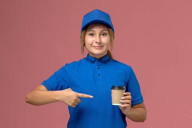 ピンクの壁にコーヒーの配達カップをポーズして保持している青い制服を着た若い女性の宅配便の正面図、サービス制服配達女性の仕事