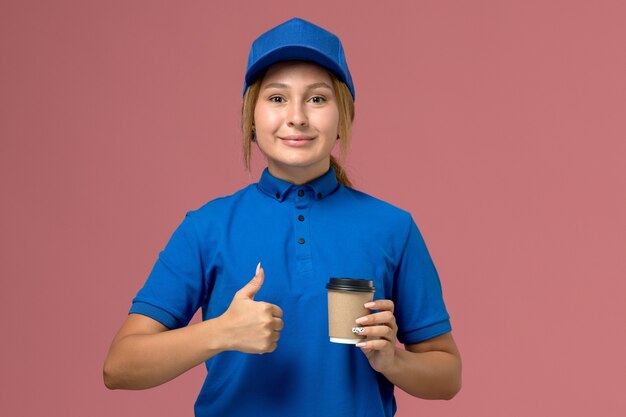 ピンクの壁にコーヒーの配達カップをポーズして保持している青い制服の正面図若い女性の宅配便、サービス制服配達女性