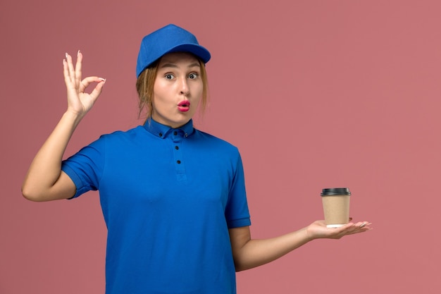 Front view young female courier in blue uniform posing holding delivery cup of coffee, service job uniform delivery woman