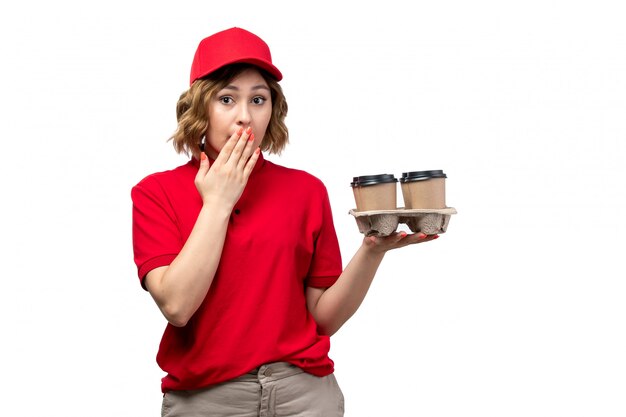 A front view young female courier female worker of food delivery service holding coffee cups with surprised expression on white