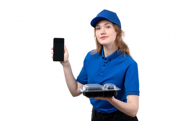 A front view young female courier female worker of food delivery service holding food bowl and using a phone with smile on white